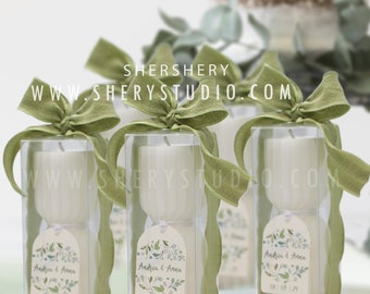 Puede incluir: Tres pequeñas velas blancas en cajas transparentes con lazos de cinta verde y etiquetas. Las etiquetas tienen un diseño de acuarela de hojas verdes y el texto "Andrea & Anna 08.09.24".
