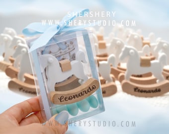 Puede incluir: Un caballo de madera blanco con un lazo azul y una caja de plástico transparente llena de caramelos azules y blancos. La caja tiene el texto "Leonardo" y el sitio web "www.sherystudio.com".