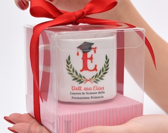 Puede incluir: Una taza de cerámica blanca dentro de una caja de regalo de plástico transparente con un lazo rojo. La taza presenta una letra E roja con un birrete de graduación, rodeada de hojas de laurel verdes y el texto Dott.ssa Elisa Laurea in Scienze della Formazione Primaria.