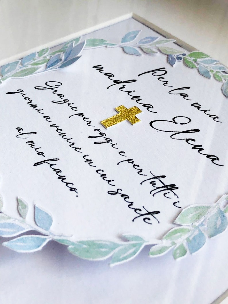 Puede incluir: Una tarjeta blanca con una cruz dorada y una corona de laurel acuarela azul y verde. La tarjeta dice "Per la mia madrina Elena Grazie per oggi e per tutti i giorni a venire in cui sarai al mio fianco."