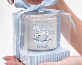 Puede incluir: Una taza de cerámica blanca con una ilustración de un caballo balancín azul y el texto Battesimo di Noah, presentada en una caja de regalo cuadrada transparente con una cinta azul claro y base a rayas como objeto decorativo.
