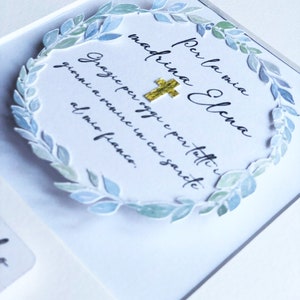 Puede incluir: Una tarjeta blanca con una corona de acuarela azul y verde que rodea el texto "Per la mia madrina Elena" y "Grazie per oggi e per tutti i giorni a venire in cui sarete al mio fianco."