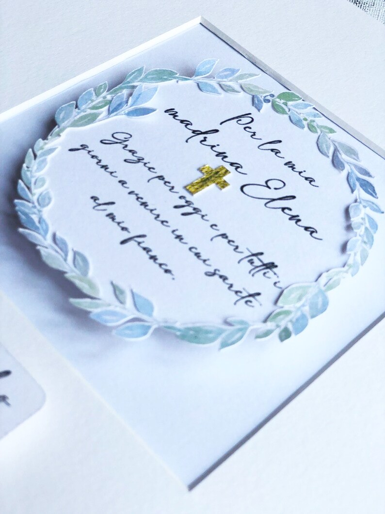Puede incluir: Una tarjeta blanca con una corona de acuarela azul y verde que rodea el texto "Per la mia madrina Elena" y "Grazie per oggi e per tutti i giorni a venire in cui sarete al mio fianco."
