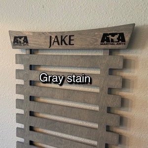 May include: Gray stained wooden belt rack with 10 slots for belts. The rack has a top piece with the text "JAKE" and the logo "AZA MARTIAL ARTS" on each side.