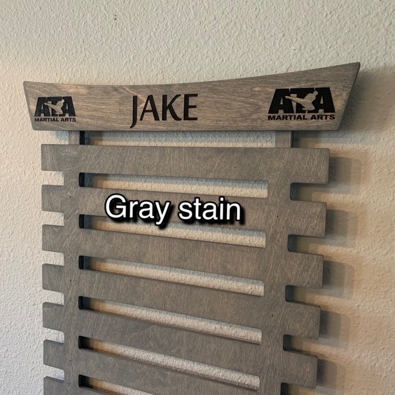 May include: Gray stained wooden belt rack with 10 slots for belts. The rack has a top piece with the text "JAKE" and the logo "AZA MARTIAL ARTS" on each side.
