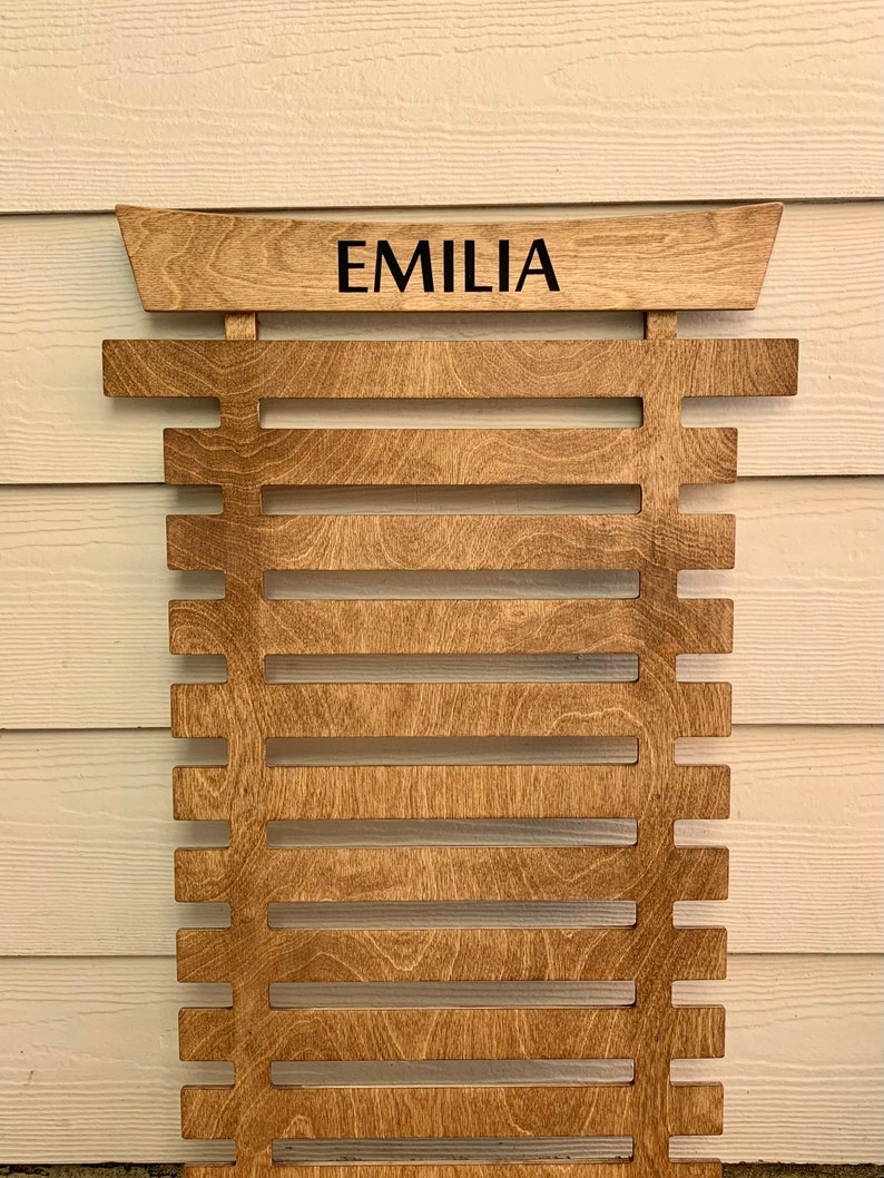 May include: A wooden wall-mounted belt rack with the name "EMILIA" engraved on the top. The rack has 12 slots for belts.
