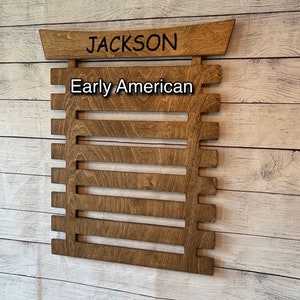 May include: A wooden wall-mounted belt rack with the name "JACKSON" and the words "Early American" written on it. The rack has eight slots for belts.