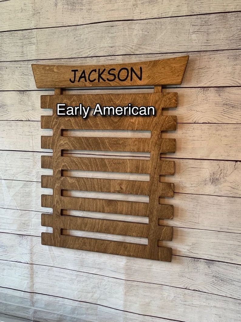 May include: A wooden wall-mounted belt rack with the name "JACKSON" and the words "Early American" written on it. The rack has eight slots for belts.