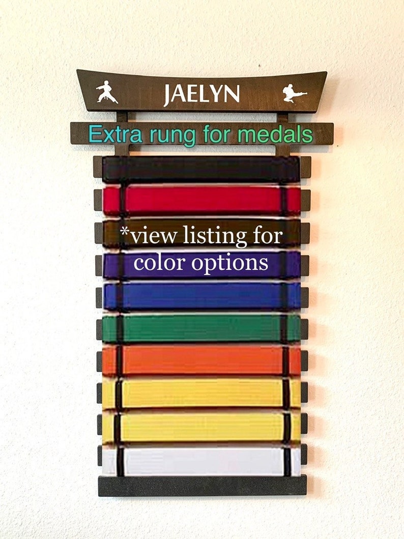 May include: A wooden martial arts belt rack with the name "Jaelyn" and the text "Extra rung for medals" on it. The rack has 9 slots for belts and an extra rung for medals. The text "*view listing for color options" is also on the rack.