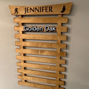 May include: A wooden wall-mounted display rack for martial arts belts. It has a curved top with the name "JENNIFER" and the words "Golden Oak" printed on the front.