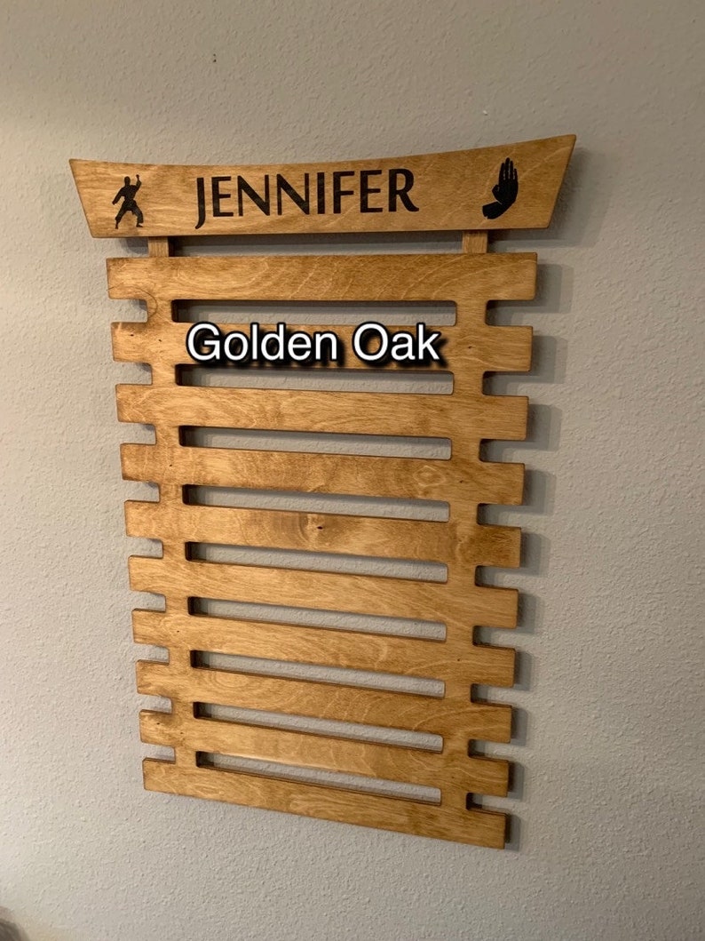 May include: A wooden wall-mounted display rack for martial arts belts. It has a curved top with the name "JENNIFER" and the words "Golden Oak" printed on the front.