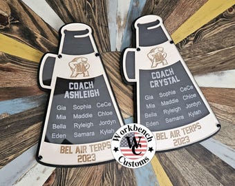 May include: Two black megaphone-shaped wooden signs with gold lettering. The signs are decorated with a turtle logo and the text "Coach Ashleigh" and "Coach Crystal". The signs list the names of players on a team, including Gia, Sophia, Mia, Maddie, Bella, Ryleigh, Eden, Samara, and Kylah. The signs also include the text "Bel Air Terps 2023".