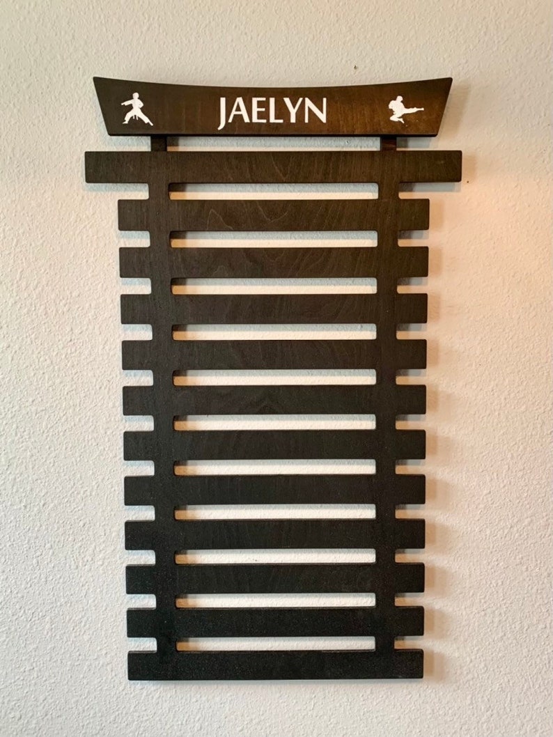 May include: A dark brown wooden martial arts belt rack with the name "JAELYN" printed on the top. The rack has 12 slots for belts and features a karate silhouette on each side.