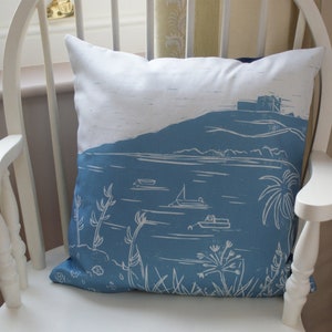 May include: A square decorative pillow with a blue and white landscape design. The pillow features a coastal scene with boats, plants, and a building on a hill. The pillow is resting on a white wooden chair.