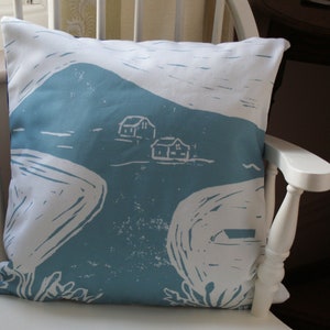 May include: Square throw pillow with a blue and white landscape design. The pillow features a coastal scene with two houses, a body of water, and a shoreline. The pillow is placed on a white wooden chair.