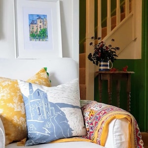 Peut inclure: Intérieur de salon avec un canapé blanc orné de coussins décoratifs et d'un plaid coloré. Une peinture encadrée est accrochée au mur. Une petite table en bois supporte un vase de fleurs. L'ambiance est lumineuse et accueillante.