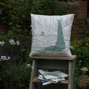 May include: A white throw pillow with a green print of a lighthouse and a sailboat on a body of water. The pillow is sitting on a wooden stool with a white and green striped fabric on the shelf below.