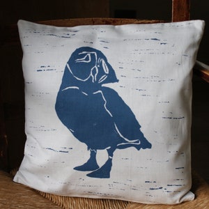 May include: A white pillow with a blue silhouette of a puffin printed on it. The background of the pillow has a subtle blue and white striped pattern.