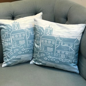 May include: Two blue throw pillows with a white cityscape design. The pillows feature a simple, graphic design of buildings with a clock tower.