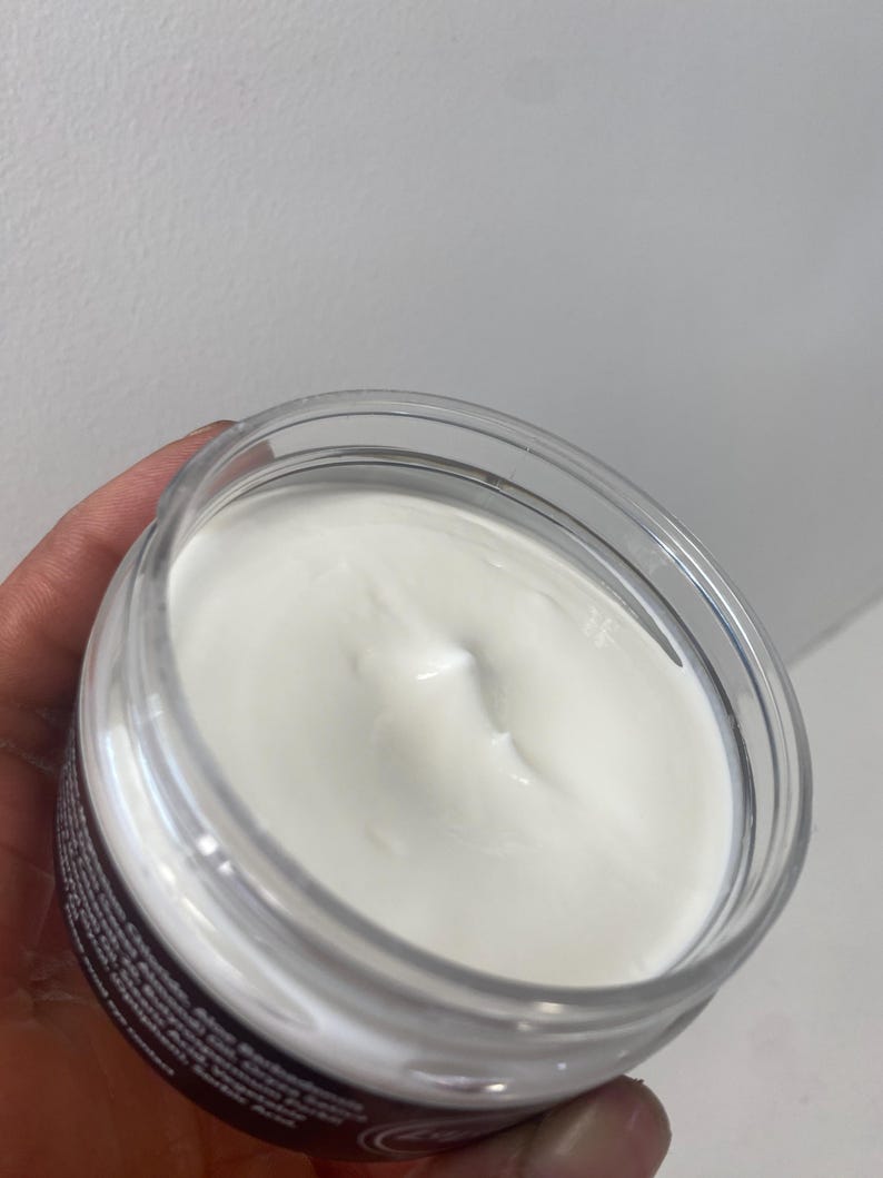 May include: A clear, round jar filled with a white, creamy substance. The jar has a clear lid and a dark label around the base. The cream has a smooth, slightly textured surface.