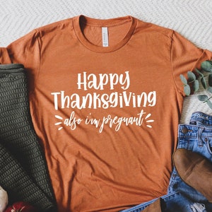 May include: A burnt orange t-shirt with the text "Happy Thanksgiving also I'm pregnant" in white lettering. The shirt is laid out on a white and grey chevron patterned surface with a green sweater, brown boots, and blue jeans.