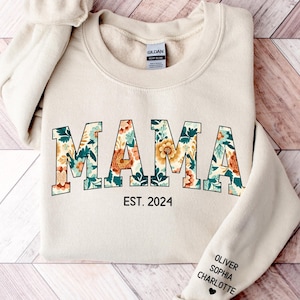 Op de afbeelding: Een crèmekleurige sweatshirt met het woord "MAMA" in een bloemenpatroon. Onder het woord "MAMA" staat de tekst "EST. 2024". Op de mouw staan de namen "OLIVER", "SOPHIA" en "CHARLOTTE".