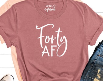 May include: A dusty rose t-shirt with the text "Forty AF" in white script. The shirt is laid out on a white wood background with a pair of light grey trainers and a pair of blue jeans.