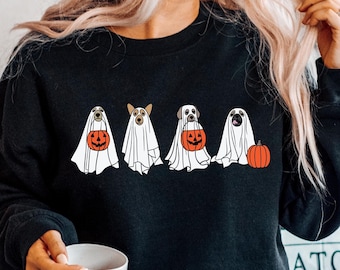 Puede incluir: Una sudadera negra con un gráfico de cuatro perros vestidos de fantasmas sosteniendo calabazas. Los perros llevan trajes de fantasmas blancos con calabazas naranjas en sus patas. La sudadera la lleva una persona con el pelo largo y rubio.