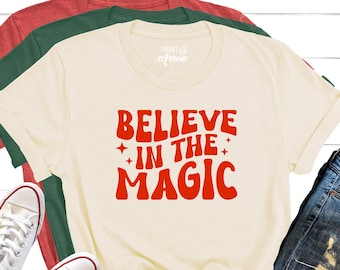 May include: Cream-colored t-shirt with the words "BELIEVE IN THE MAGIC" in red, paired with blue jeans and white sneakers. Additional t-shirts in red and green are visible. The image is styled on a white wooden background.