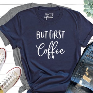 May include: A navy blue t-shirt with the text "But First Coffee" in white lettering. The shirt is laid out on a white wooden surface with a pair of white trainers and a pair of blue jeans.