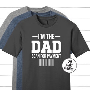以下が含まれることがあります： 白文字で「I'm the Dad Scan for Payment」と書いてあるダークグレーのTシャツ。下にバーコードがあります。このTシャツは20色展開です。