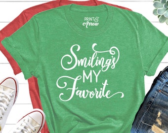 May include: Green t-shirt with white text that reads "Smiling's My Favorite". The shirt is shown on a white wooden background with a pair of white trainers and a pair of blue jeans.