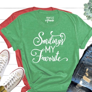May include: Green t-shirt with white text that reads "Smiling's My Favorite". The shirt is shown on a white wooden background with a pair of white trainers and a pair of blue jeans.