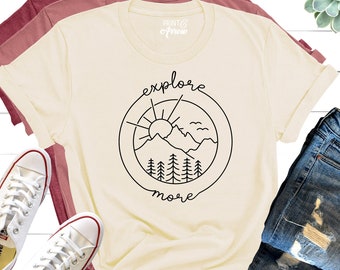 May include: A cream coloured t-shirt with a black graphic of a mountain range, sun, and birds inside a circle with the text "explore more".