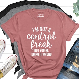 May include: A pink t-shirt with the text "I'm not a control freak but you're doing it wrong" in white lettering. The shirt is laid out on a white wooden surface with a pair of white sneakers and a pair of blue jeans.