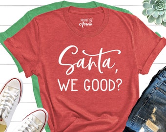 May include: Red t-shirt with white text that says "Santa, We Good?"  The shirt is laid out on a white surface with a pair of white sneakers, a pair of blue jeans, and a small green succulent plant.