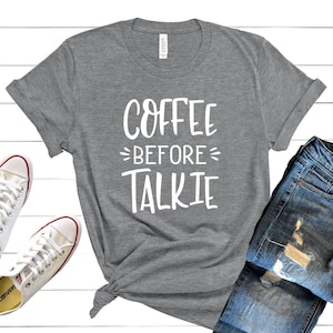 May include: A heather gray t-shirt with the text "Coffee Before Talkie" in white lettering. The shirt is laid out on a white wooden surface with a pair of white sneakers and a pair of blue jeans.