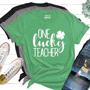Op de afbeelding: Groen T-shirt met de tekst "One lucky teacher" in wit met een wit vierbladig klavertje. Het shirt wordt getoond op een witte houten achtergrond met jeans en sneakers.