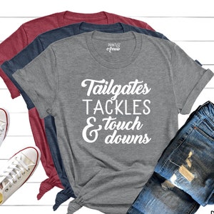 May include: Three t-shirts in maroon, navy, and heather gray with the text "Tailgates Tackles & Touchdowns" in white. The shirts are paired with blue jeans and white sneakers. The Print & Arrow logo is visible.