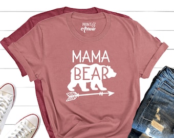 May include: A pink t-shirt with the text "Mama Bear" in white lettering and a white bear silhouette with an arrow pointing to the right. The shirt is laid out on a white wooden surface with a pair of white sneakers and a pair of blue jeans.