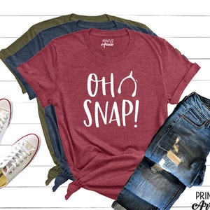 May include: A maroon t-shirt with the text "Oh Snap!" in white lettering and a white horseshoe graphic. The shirt is folded and displayed with a pair of blue jeans and two pairs of white sneakers.