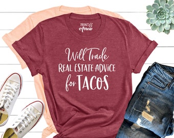 May include: A maroon t-shirt with the text "Will Trade Real Estate Advice for Tacos" in white lettering. The shirt is laid out on a white wooden surface with a pair of white sneakers, a pair of blue jeans, and a small potted succulent plant.