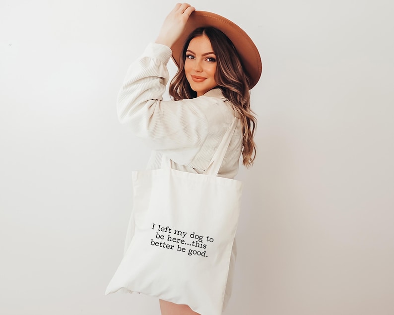 May include: Cream-colored tote bag with the text "I left my dog to be here...this better be good." The bag is carried by a person wearing a brown hat and a cream-colored shirt. The bag is made of a soft, durable material.