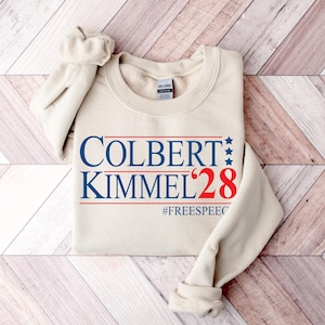 May include: A cream-colored sweatshirt with the text "COLBERT KIMMEL '28" in blue and red, with three blue stars and the hashtag "#FREESPEECH". The sweatshirt has a crew neck and long sleeves.