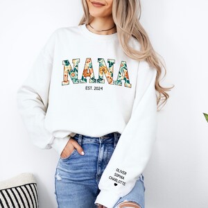 Op de afbeelding: Witte trui met het woord "NANA" in bloemenletters en de tekst "EST. 2024" eronder. De trui heeft een zwart hart en de namen "Oliver", "Sophia" en "Charlotte" gedrukt op de linkermouw.