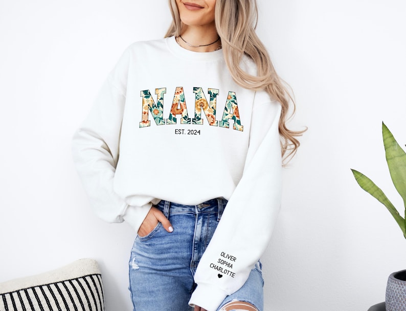 Op de afbeelding: Witte trui met het woord "NANA" in bloemenletters en de tekst "EST. 2024" eronder. De trui heeft een zwart hart en de namen "Oliver", "Sophia" en "Charlotte" gedrukt op de linkermouw.