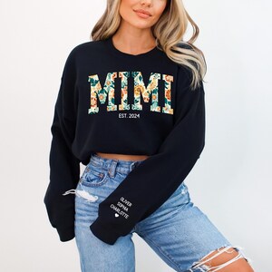 May include: Black cropped sweatshirt with the word "MIMI" in floral print and the text "EST. 2024" below. The sweatshirt has long sleeves and a white heart with the names "OLIVER, SOPHIA, CHARLOTTE" on the left sleeve.