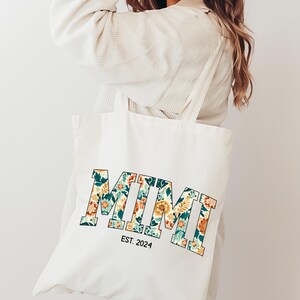 May include: A white canvas tote bag with a floral print design that spells out "MIMI" in bold letters. The bag also has the text "EST. 2024" printed below the floral design.