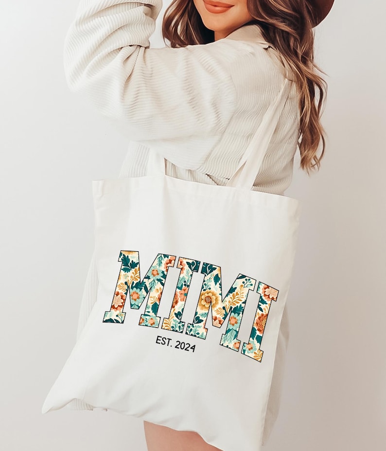 May include: A white canvas tote bag with a floral print design that spells out "MIMI" in bold letters. The bag also has the text "EST. 2024" printed below the floral design.