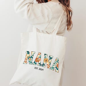 Op de afbeelding: Witte canvas draagtas met het woord "NANA" in grote, gedurfde letters. De letters zijn gevuld met een bloemenpatroon in tinten oranje, geel, groen en blauw. De tekst "EST. 2024" is onder het woord "NANA" gedrukt.